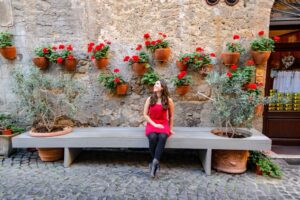Kate Storm sitting on a bench in Orvieto Italy. She's wearing a red dress, black tights, and black boots. There are red flowers in pots on the wall behind her.
